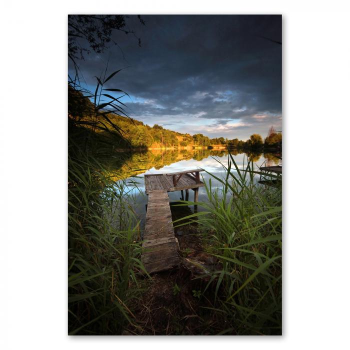 Holzsteg durch Schilfgras führt zu Plattform auf ruhigem See, Himmel strahlt in warmen Farben des Sonnenuntergangs.