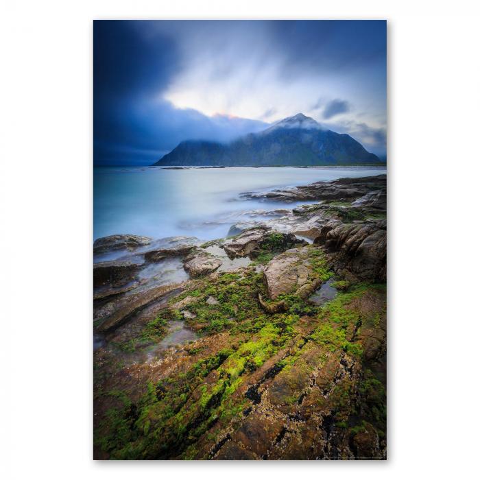 Dramatische Wolken hängen über einem Berg am Fjord.Im Vordergrund der Küste liegen von Algen bewachsene Felsen.