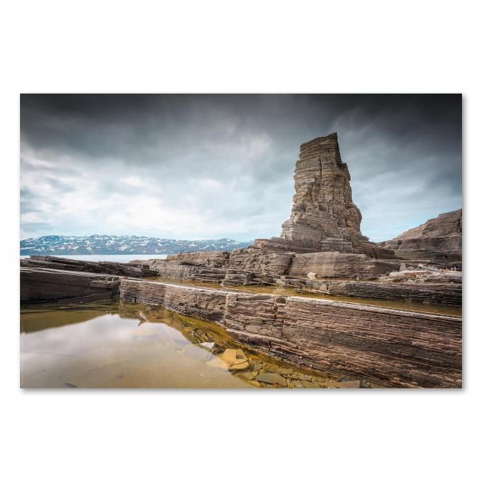 Ein hoher, schmaler Felsen ragt an der norwegischen Küste aus dem Wasser, während sich der Himmel dramatisch bewölkt.