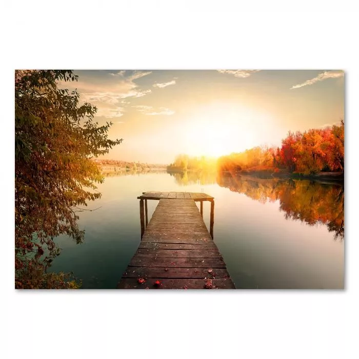 Ein Steg führt auf einen See hinaus. Herbstliche Bäume säumen das Ufer & spiegeln sich im Wasser. Die Sonne taucht die Landschaft in warme Farben.