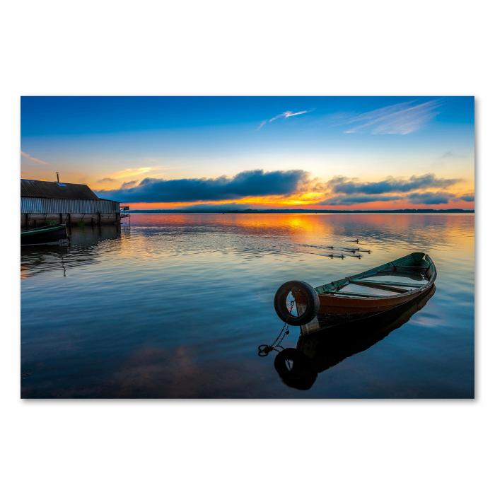 Altes Holzboot am Seeufer, Sonnenaufgang hinter Wolken, warme Farben am Himmel.