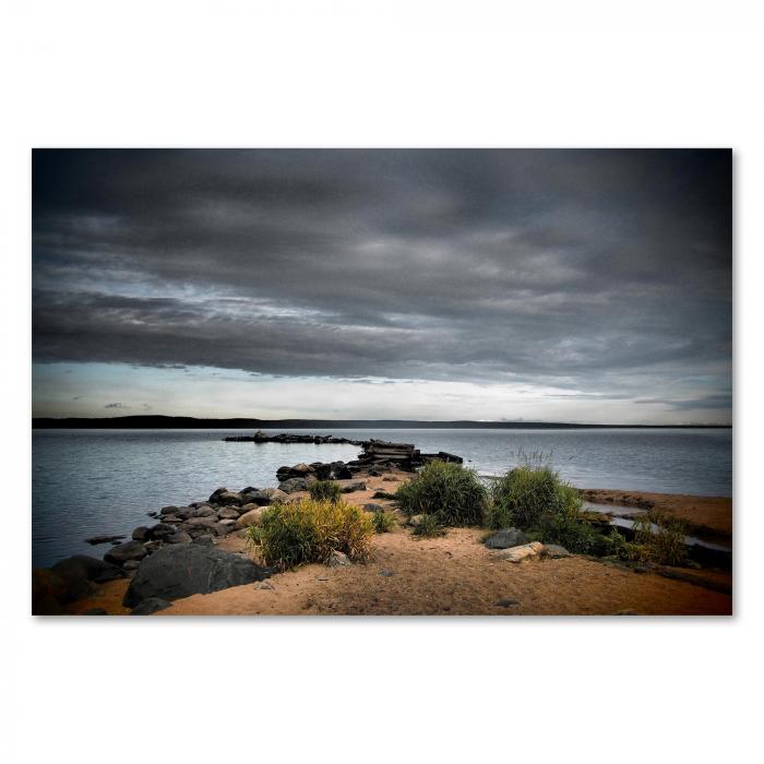 Blick über einen See mit Steinen und Gras im Vordergrund und dunklen Wolken am Himmel.