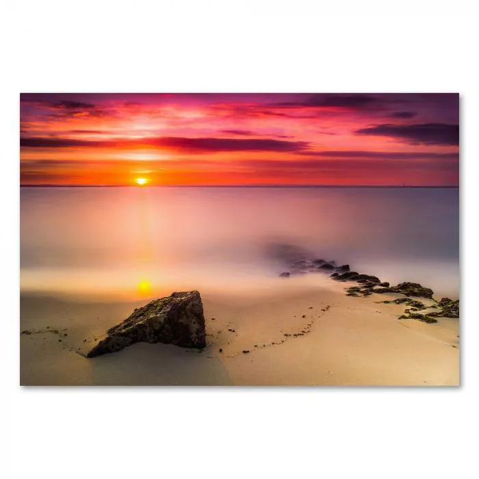 Ein Felsen am Strand im Vordergrund, im Hintergrund die untergehende Sonne über dem Meer.