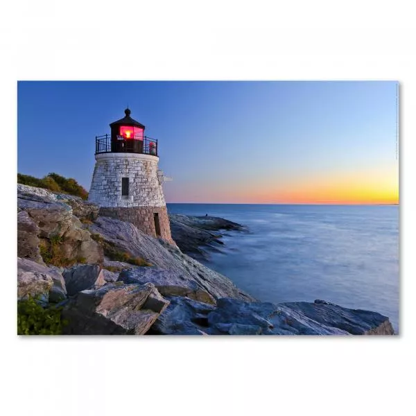 Ein weißer Leuchtturm steht auf felsiger Küste vor einem blauen Meer und einem orange-roten Abendhimmel.