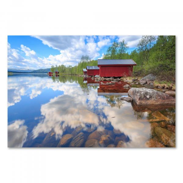 Zwei rote Holzhütten stehen am Ufer eines Sees.Der blaue Himmel mit weißen Wolken spiegelt sich im Wasser.