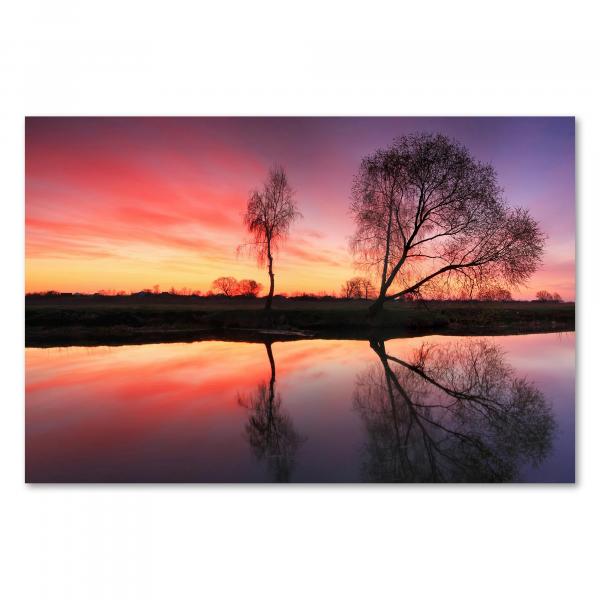 Bäume spiegeln sich im Wasser, der Himmel erstrahlt in leuchtenden Farben – ein farbenfroher Sonnenaufgang am Fluss.